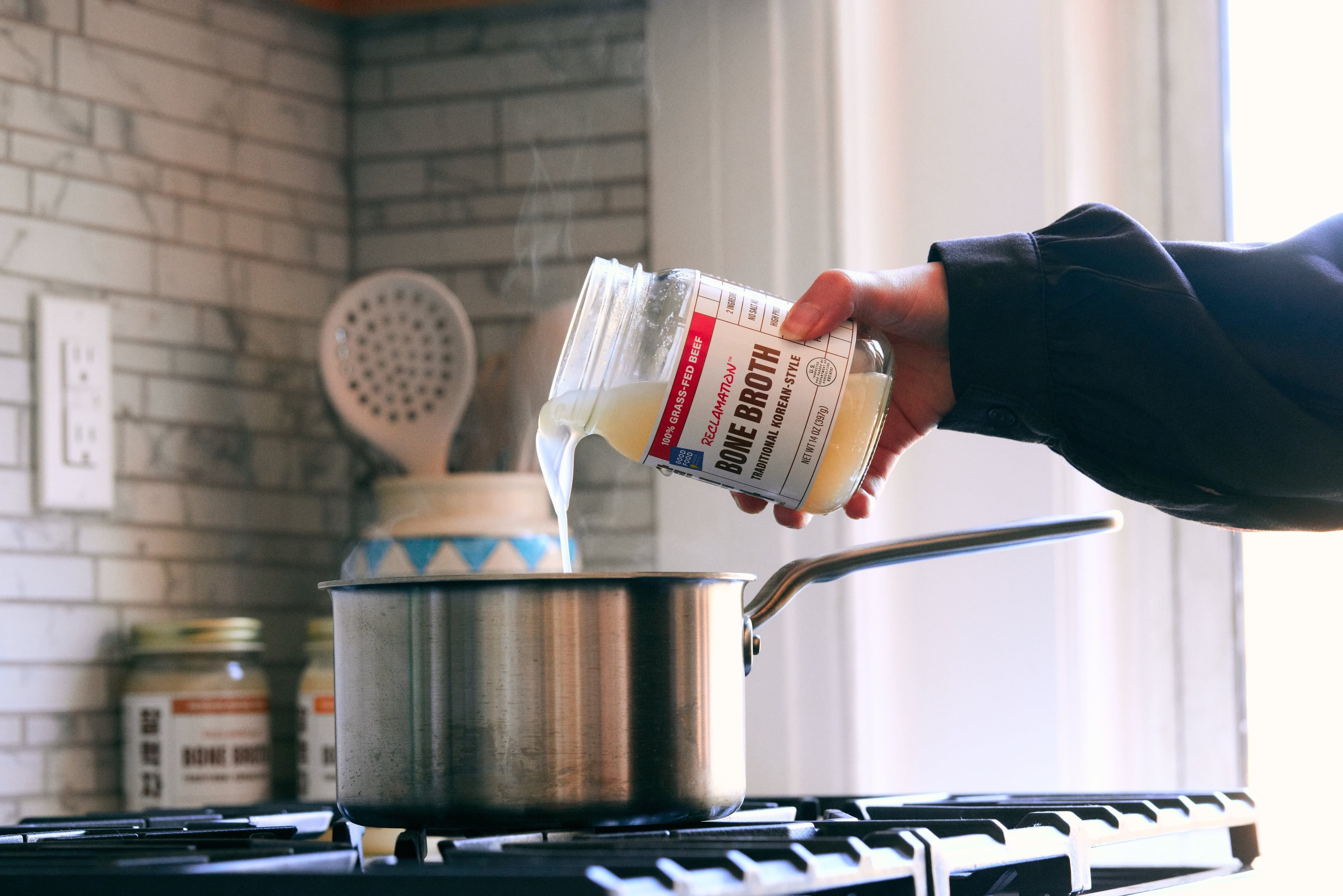 Person pouring Reclamation Korean-Style Beef Bone Broth from a jar into a pot on a stove.